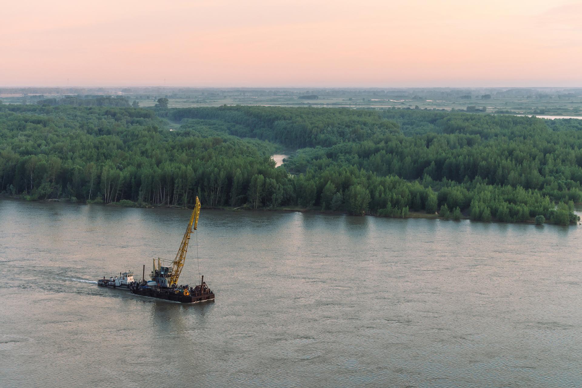 Beautiful river landscape with green shores and ship with copy space. Towboat tows barge with crane along riverbank. Dawn reflexed on calm glare water surface. Pink sunset sky on background.